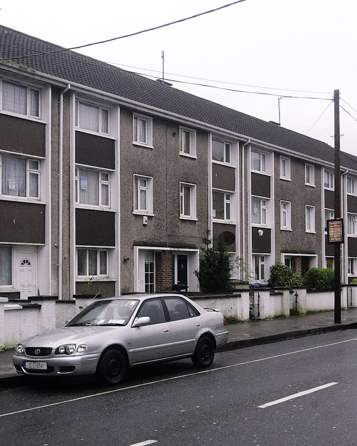 a terraced city house, with three storeys, probably designed in the 1960s. Cars are parked on the road in front.