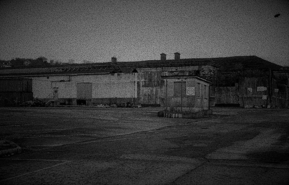 an old warehouse, boarded up with metal and wooden sheets. A security hut in the centre of the untended parking area. Black and white.