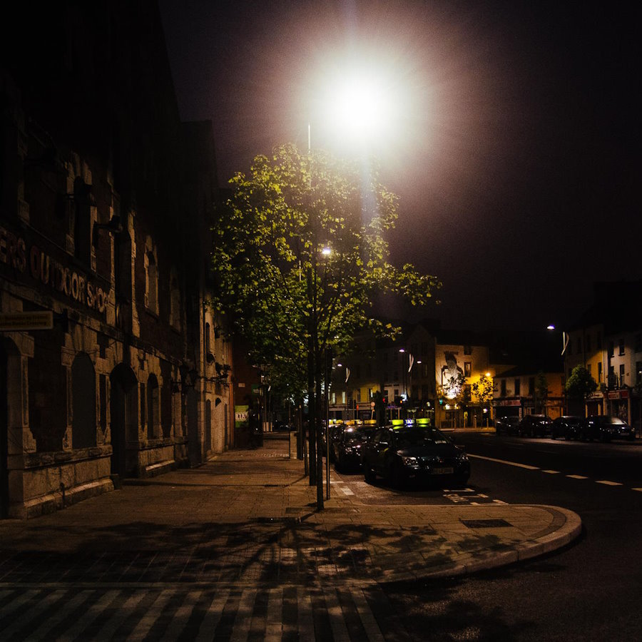 a quiet, wide city street at night with lampposts taxis and trees